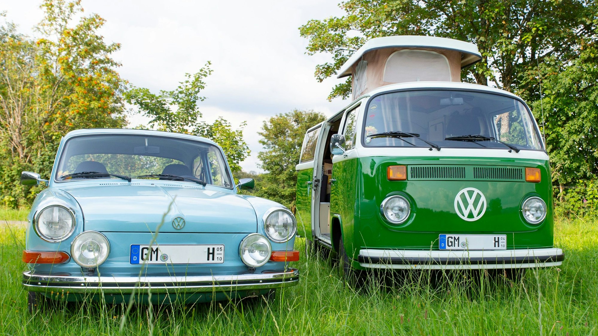 Auf einer Wiese stehen ein dunkelgrüner VW-Bulli T2b, Baujahr 1978, und ein VW 1600, Baujahr 1973, in hellblau nebeneinander.