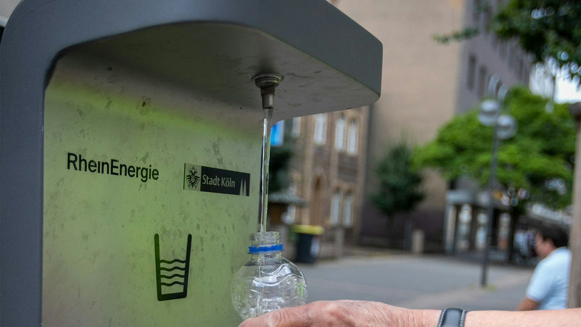 Eine Person füllt sich Wasser vom Trinkwasserbrunnen in der Porzer Innenstadt in eine Wasserflasche.