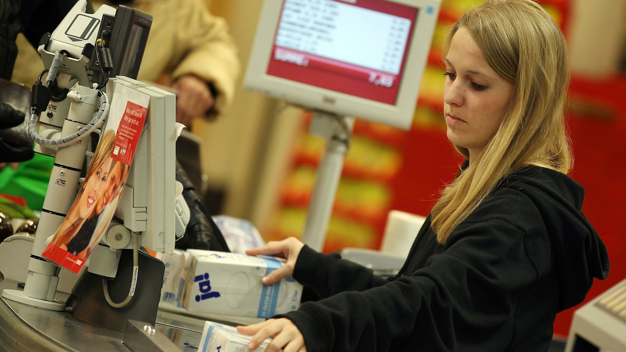 Eine junge blonde Kassiererin sitzt an einer Kasse und scannt Lebensmittel ein, die Kunden aufs Band gelegt haben. Ein Display im Hintergrund zeigt die eingescannten Preise an.