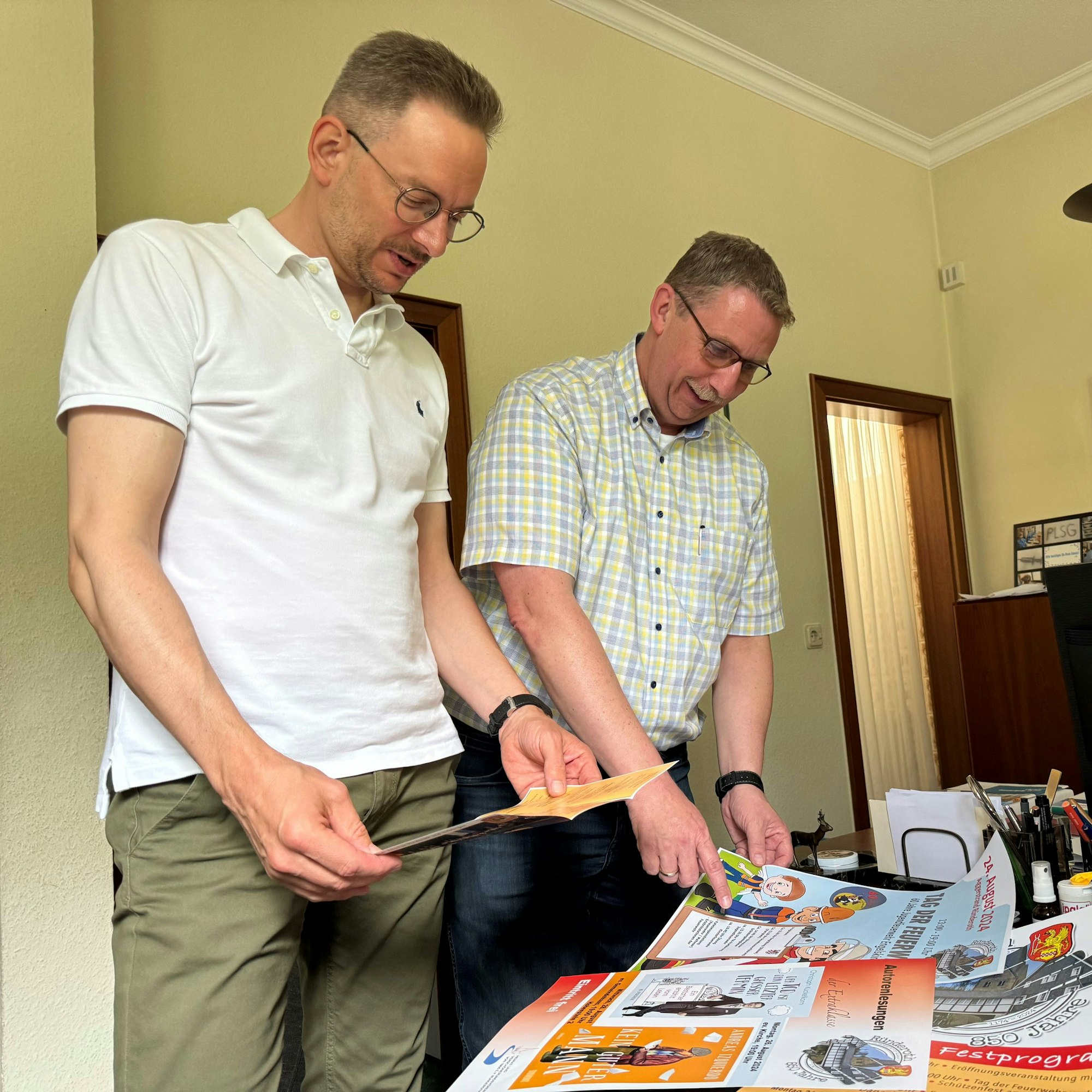 Zwei Männer stehen an einem Schreibtisch und schauen sich Plakate an, die auf Veranstaltungen während der Festwoche in Ründeroth hinweisen.