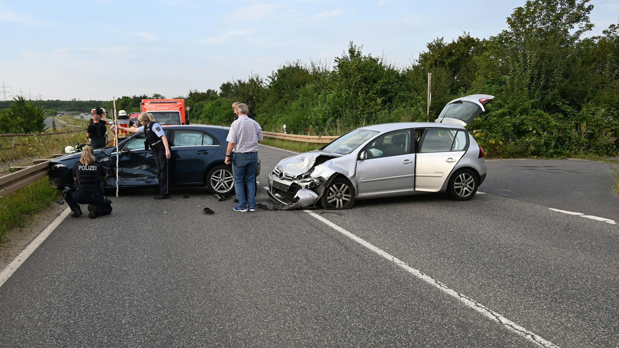 Zwei beschädigte Fahrzeuge stehen quer auf einer Straße. Auf der Fahrbahn stehen Personen, unter anderem Einsatzkräfte der Polizei. Im Hintergrund ein Rettungsfahrzeug.