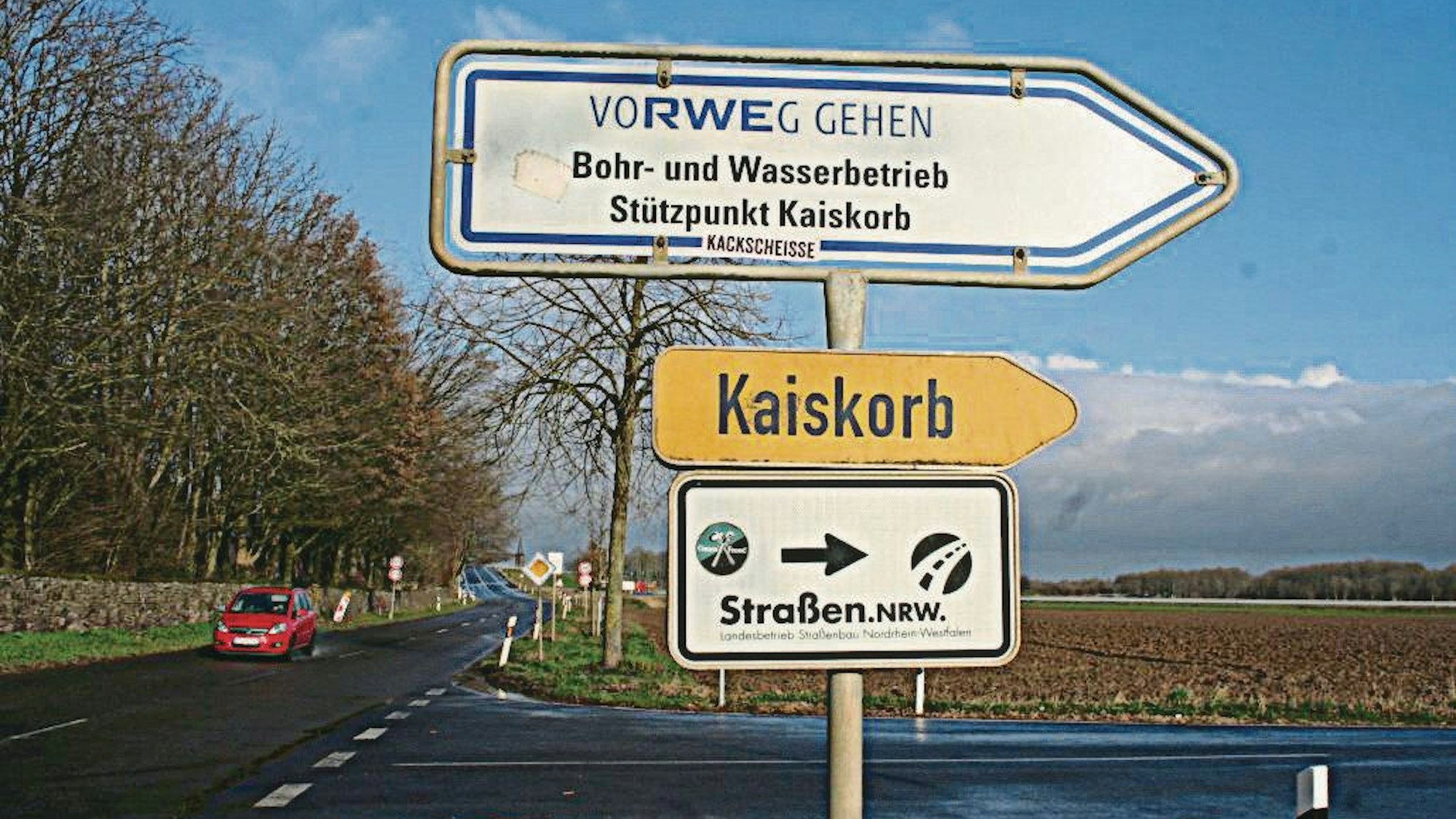 In Kaiskorb haben Straßen NRW und RWE Standorte. Auf drei Straßenschildern sind Kaiskorb, Straßen NRW und RWE abgebildet. Im Hintergrund sind Autos auf der Straße neben Feldern zu sehen.