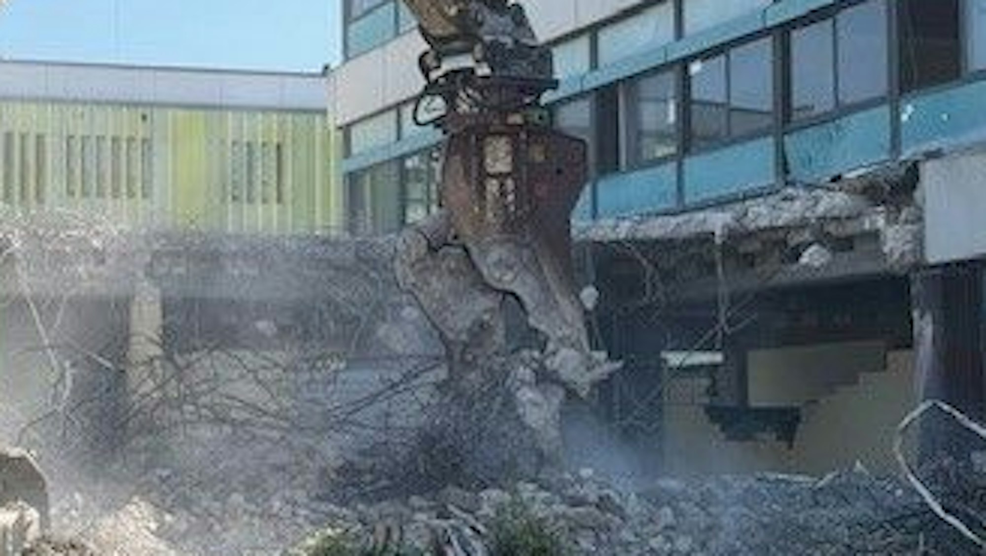 Eine Baggerzange bricht Stücke aus einem 70er-Jahre-Gebäude.