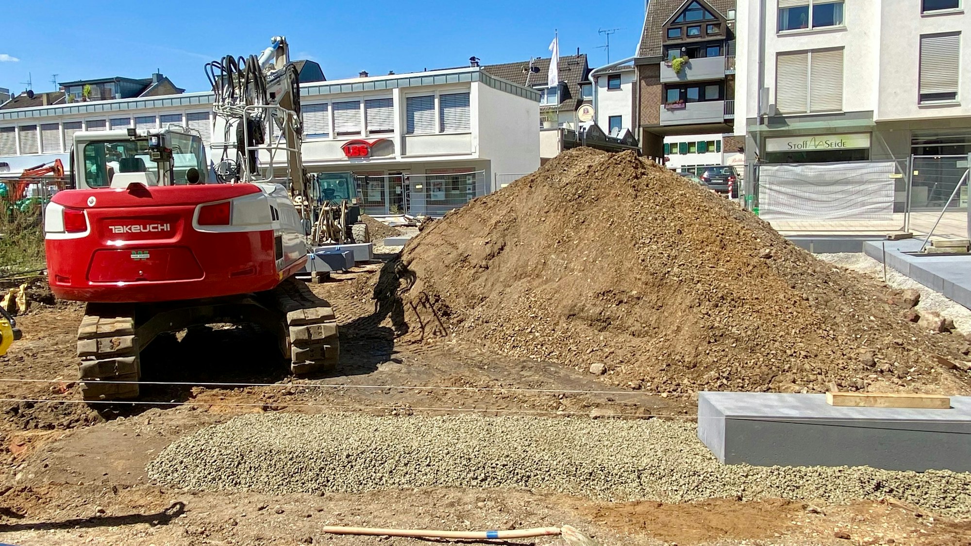 Ein Bagger neben einem Sandhaufen, im Hintergrund Häuser.