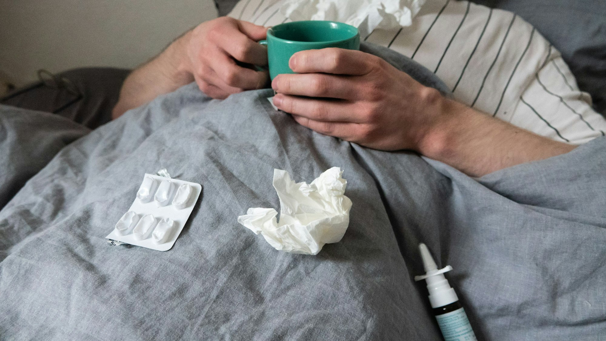 ILLUSTRATION - Ein Mann liegt mit Taschentüchern, Teetasse, Nasenspray und Tabletten im Bett (gestellte Szene). Derzeit bewegt sich die Rate akuter Atemwegserkrankungen in der Bevölkerung weiter auf einem relativ hohen Niveau im Vergleich zu früheren Jahren. +++ dpa-Bildfunk +++