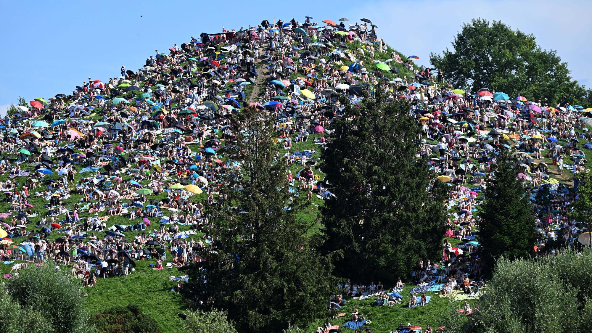 Fans sitzen auf dem Olympiaberg im Olympiapark und warten bei Sonnenschein auf den Beginn des Taylor Swift Konzerts.