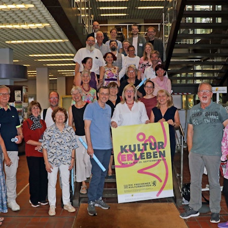 Viele Männer und Frauen lächeln in die Kamera. Zwei Frauen halten ein Schild mit der Aufschrift „KULTURERLEBEN “in ihren Händen.