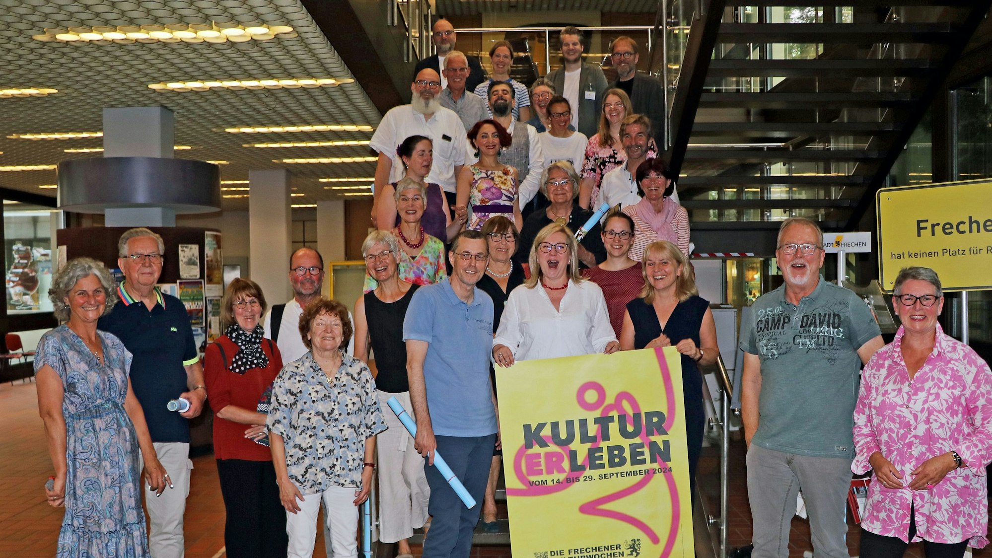 Viele Männer und Frauen lächeln in die Kamera. Zwei Frauen halten ein Schild mit der Aufschrift „KULTURERLEBEN “in ihren Händen.