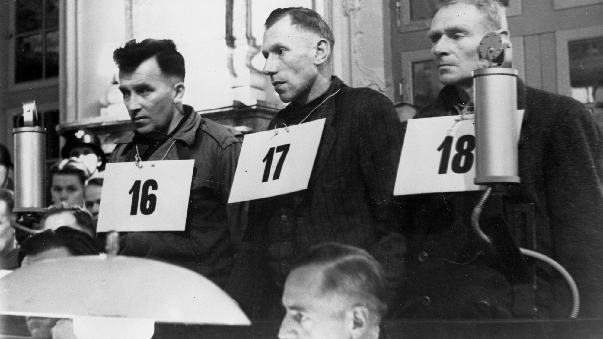 Drei Männer mit nummerierten Schildern um den Hals auf der Anklagebank.