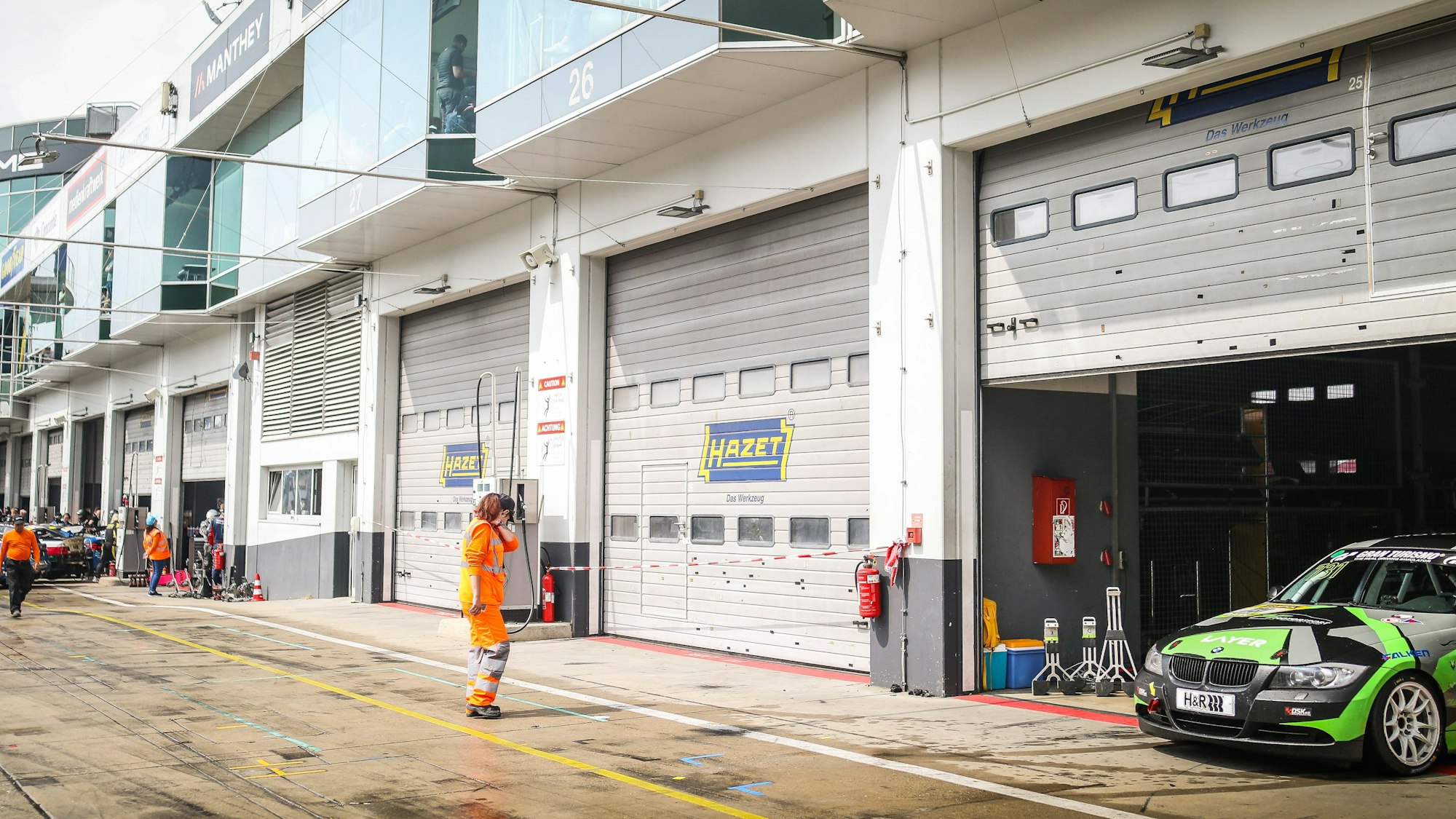 In der Boxengasse des Nürburgrings sind zwei Boxen mit rot-weißem Flatterband abgesperrt und geschlossen. Daneben sind Boxen geöffnet, in der rechten steht ein grün-schwarzer Rennwagen.