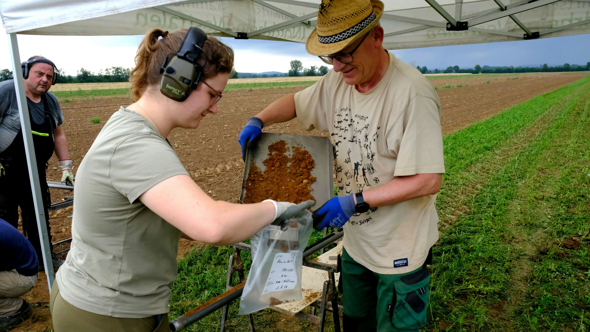Lea Garmer (trägt einen Gehörschutz) und Uwe Kalthoff von der Landwirtschaftskammer füllen eine Bodenprobe in einen Probenbeutel um. Kalthoff trägt einen Strohhut auf dem Kopf.