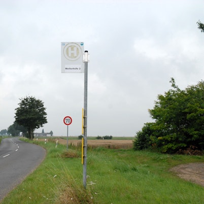 Die Bushaltestelle Mellerhöfe I steht an einer Straße. Ein Verkehrsschild zeigt Tempo 70 an. Um sie herum ist ländlicher Betrieb mit eingezäunten Weiden samt Kühen.