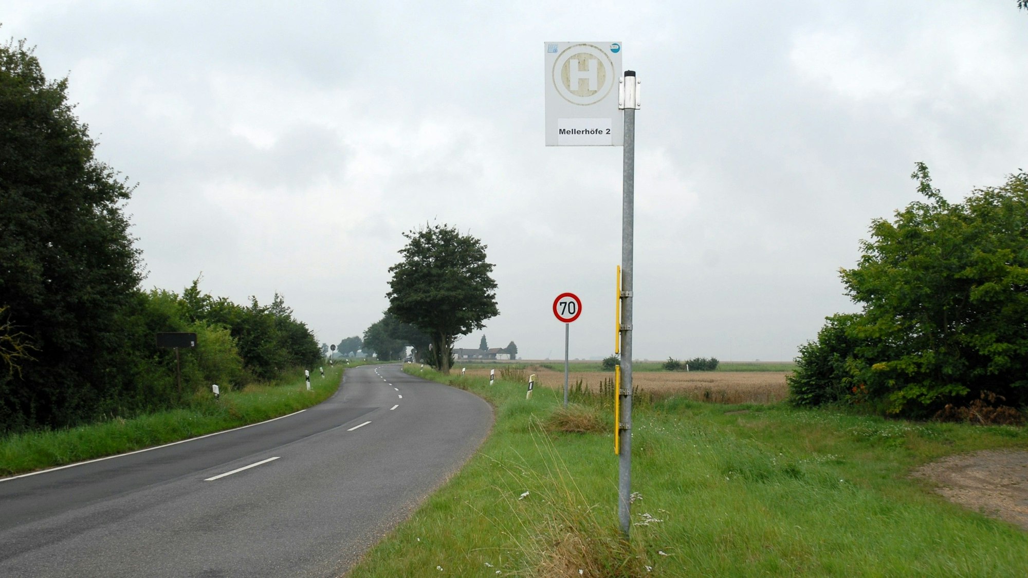 Die Bushaltestelle Mellerhöfe I steht an einer Straße. Ein Verkehrsschild zeigt Tempo 70 an. Um sie herum ist ländlicher Betrieb mit eingezäunten Weiden samt Kühen.