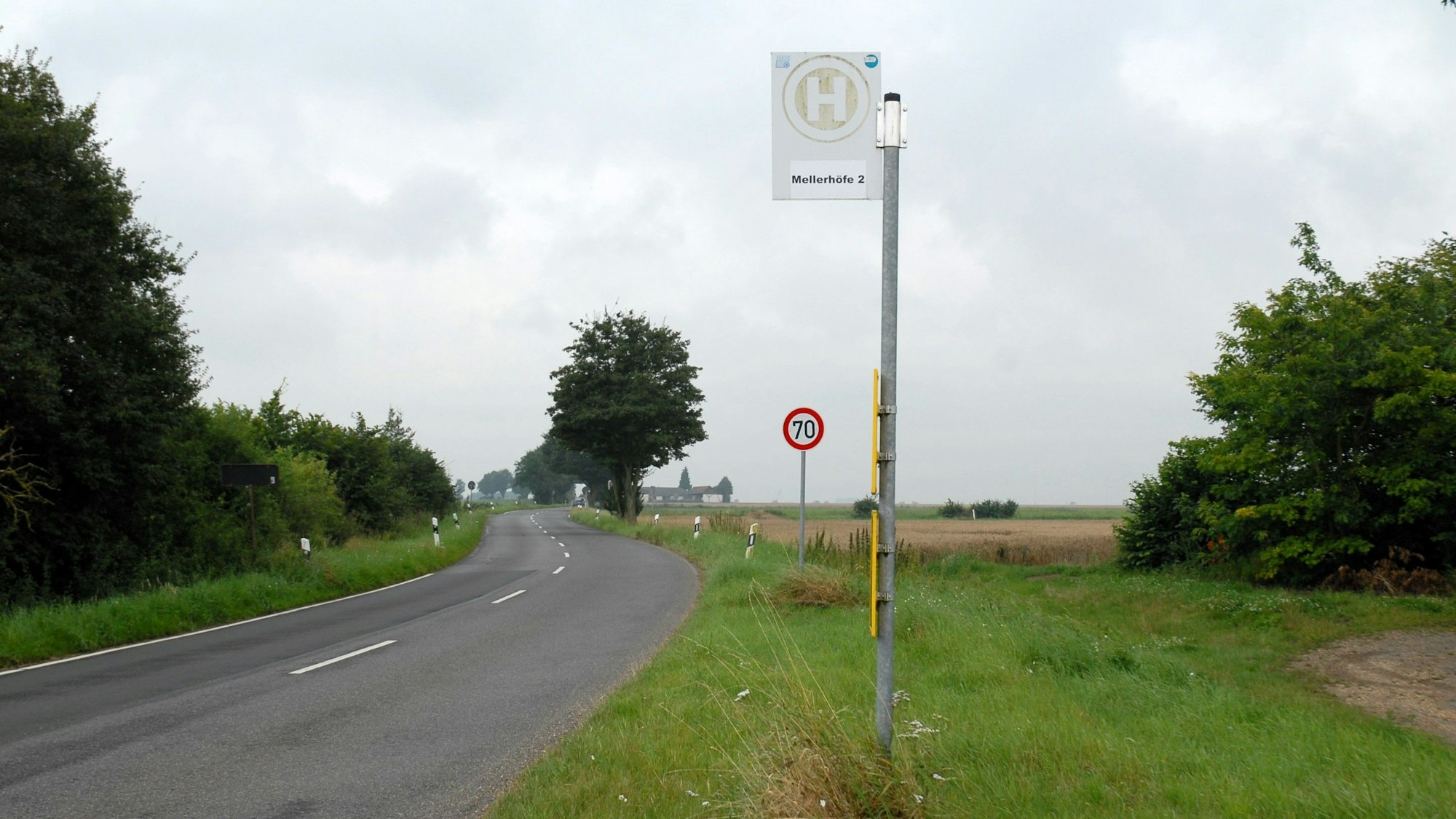 Die Bushaltestelle Mellerhöfe I steht an einer Straße. Ein Verkehrsschild zeigt Tempo 70 an. Um sie herum ist ländlicher Betrieb mit eingezäunten Weiden samt Kühen.