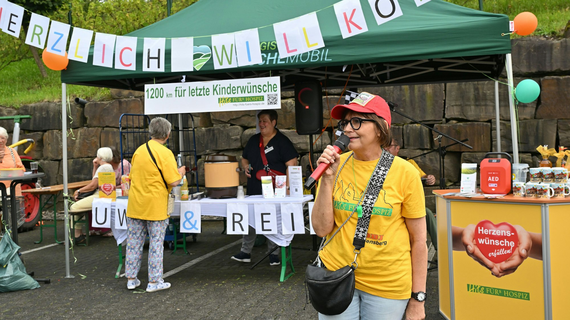 Eine Frau spricht in ein Mikro, im Hintergrund ist ein Banner mit der Aufschrift „Herzlich Willkommen“ zu sehen.