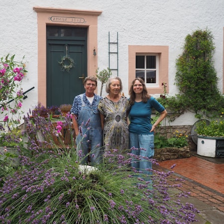 Drei Frauen, Claudia Meyer (v.l.), Renate Lorentzen und Lisa Zimmermanns, stehen in ienem Innenhof in Nettersheim, in dem viele Pflanzen wachsen. Im Hintergrund ist ein altes Haus zu sehen.