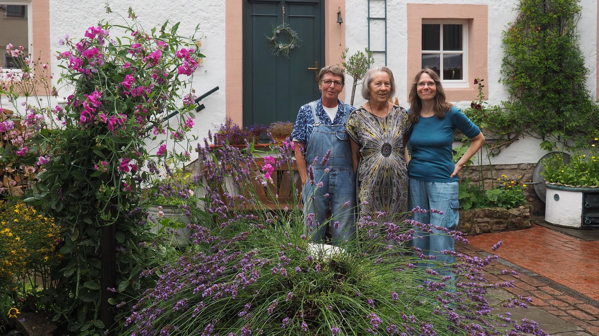 Drei Frauen, Claudia Meyer (v.l.), Renate Lorentzen und Lisa Zimmermanns, stehen in ienem Innenhof in Nettersheim, in dem viele Pflanzen wachsen. Im Hintergrund ist ein altes Haus zu sehen.