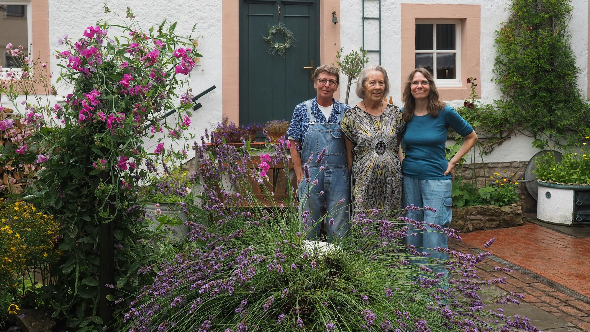 Drei Frauen, Claudia Meyer (v.l.), Renate Lorentzen und Lisa Zimmermanns, stehen in ienem Innenhof in Nettersheim, in dem viele Pflanzen wachsen. Im Hintergrund ist ein altes Haus zu sehen.