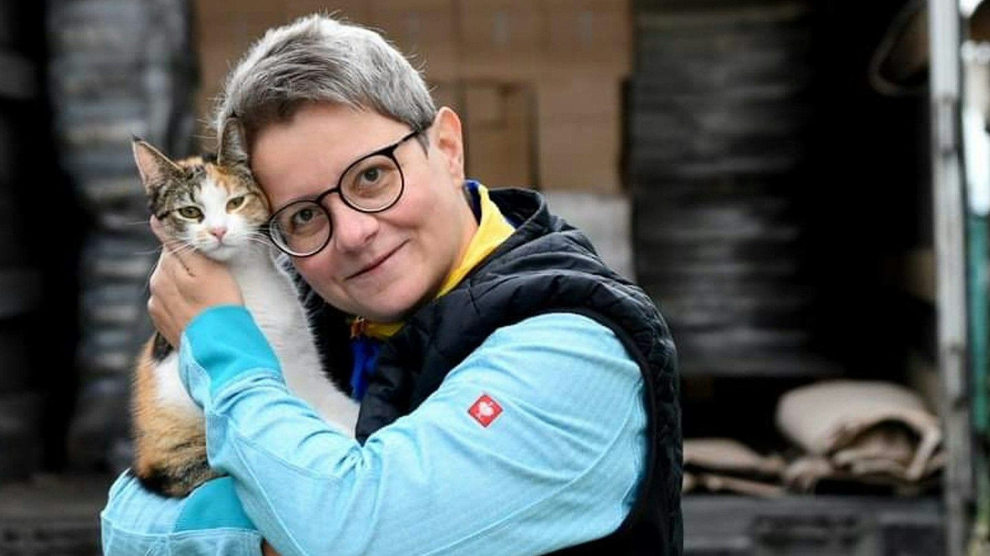 Eine Frau mit Kurzhaarschnitt, Brille und einer Katze auf dem Arm, steht vor einem beladenen Lkw.
