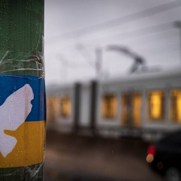 Zu sehen ist eine weiße Friedenstaube auf einer ukrainischen Flagge, die an einem Laternenmast klebt. Im Hintergrund fährt ein Stadtbahn der KVB.