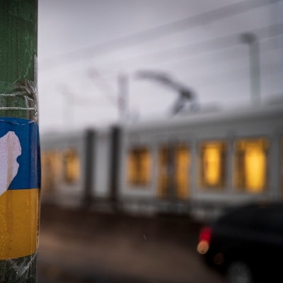 Zu sehen ist eine weiße Friedenstaube auf einer ukrainischen Flagge, die an einem Laternenmast klebt. Im Hintergrund fährt ein Stadtbahn der KVB.