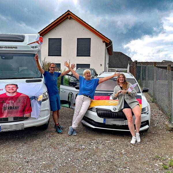 Ein Mann und zwei Frauen vor einem Wohnmobil und einem Pkw. Die Autos sind mit einem Transparent und einer Fahne geschmückt.