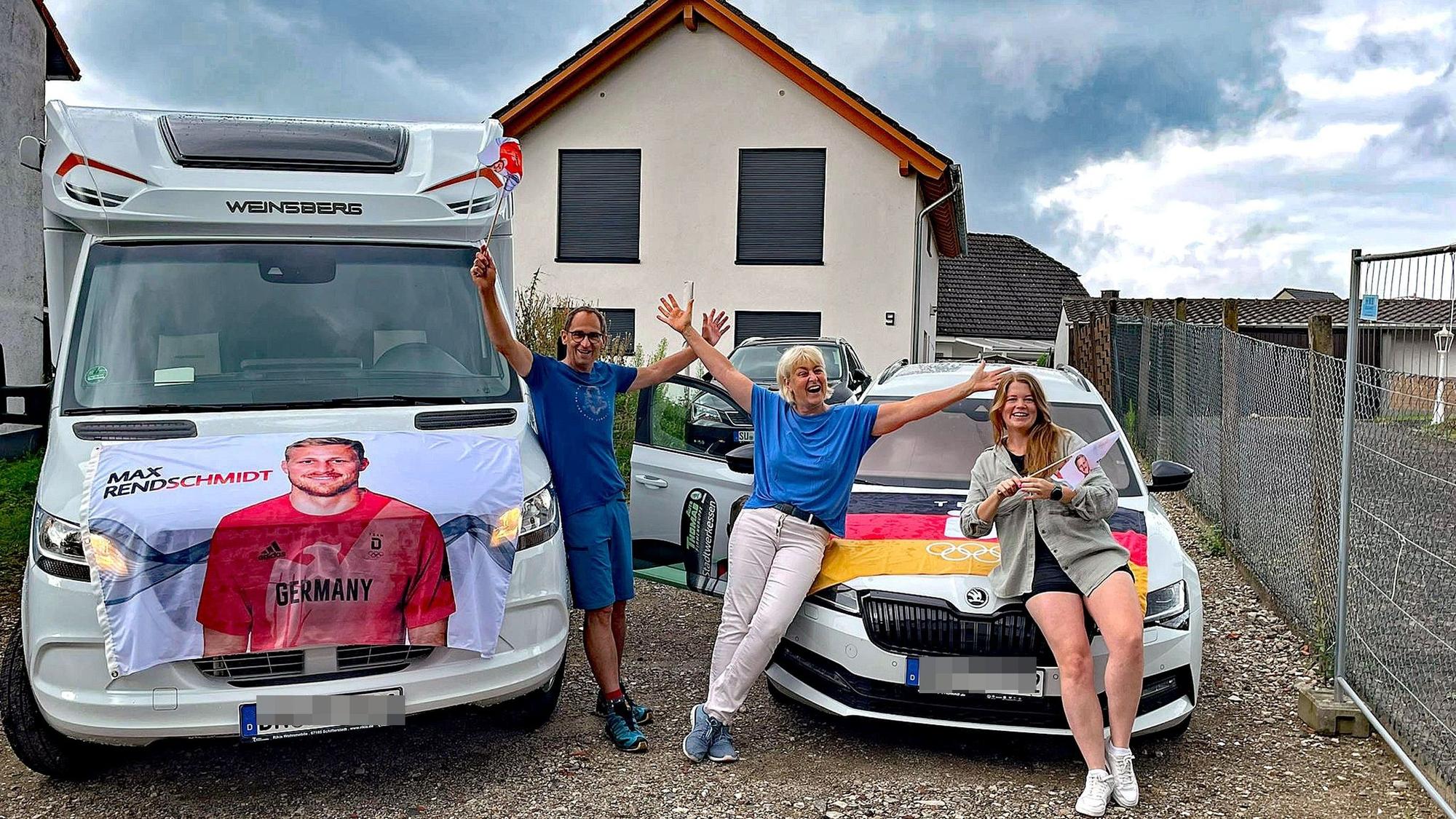 Ein Mann und zwei Frauen vor einem Wohnmobil und einem Pkw. Die Autos sind mit einem Transparent und einer Fahne geschmückt.