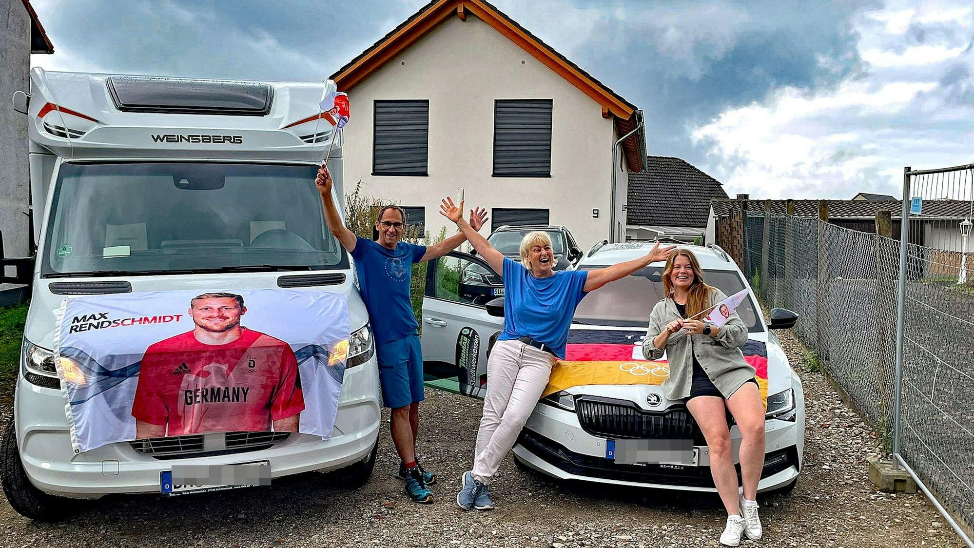 Ein Mann und zwei Frauen vor einem Wohnmobil und einem Pkw. Die Autos sind mit einem Transparent und einer Fahne geschmückt.