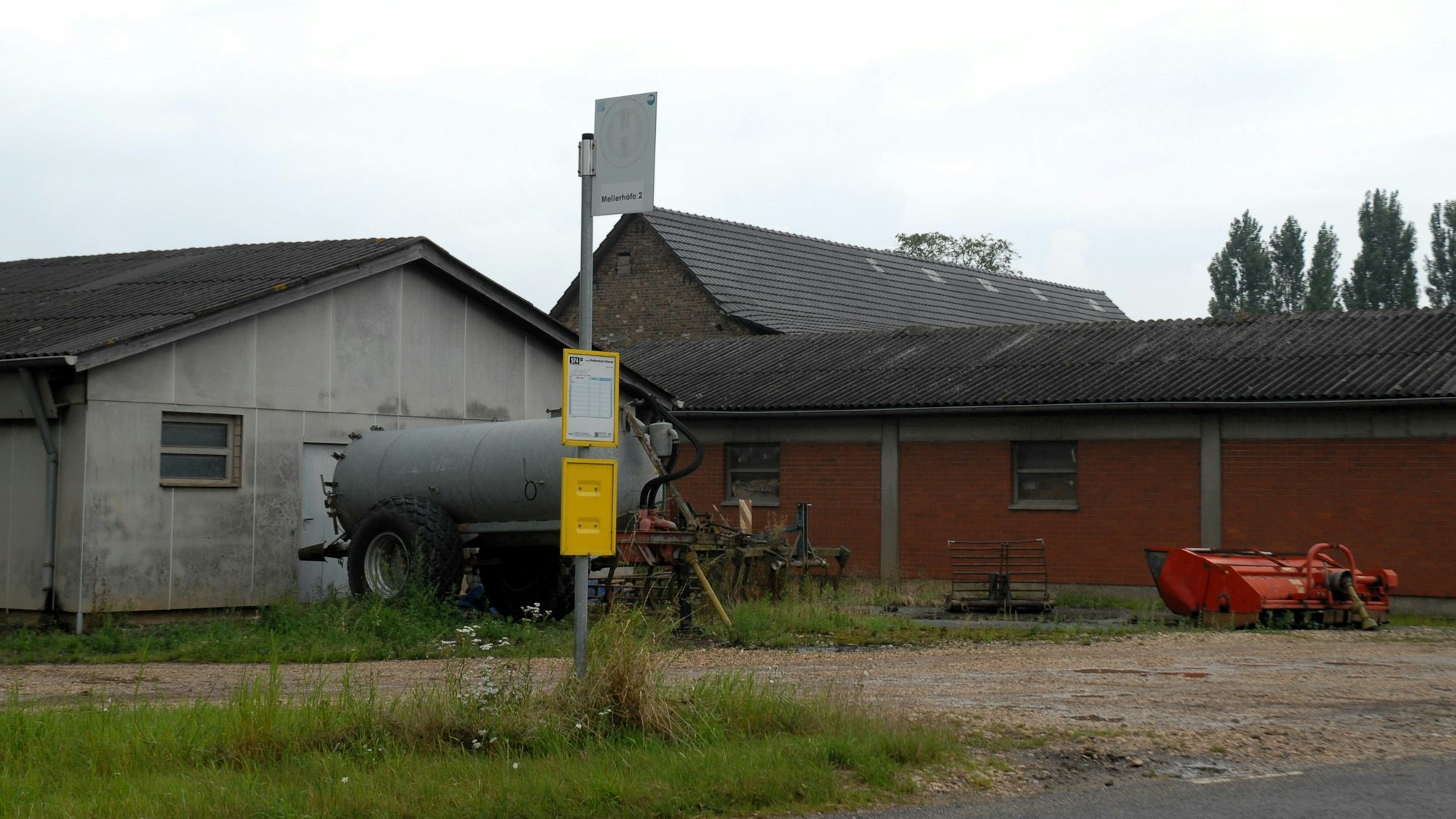 Die Bushaltestelle Mellerhöfe I steht an einer Straße vor einem Bauernhof. Um sie herum ist ländlicher Betrieb mit eingezäunten Weiden samt Kühen.