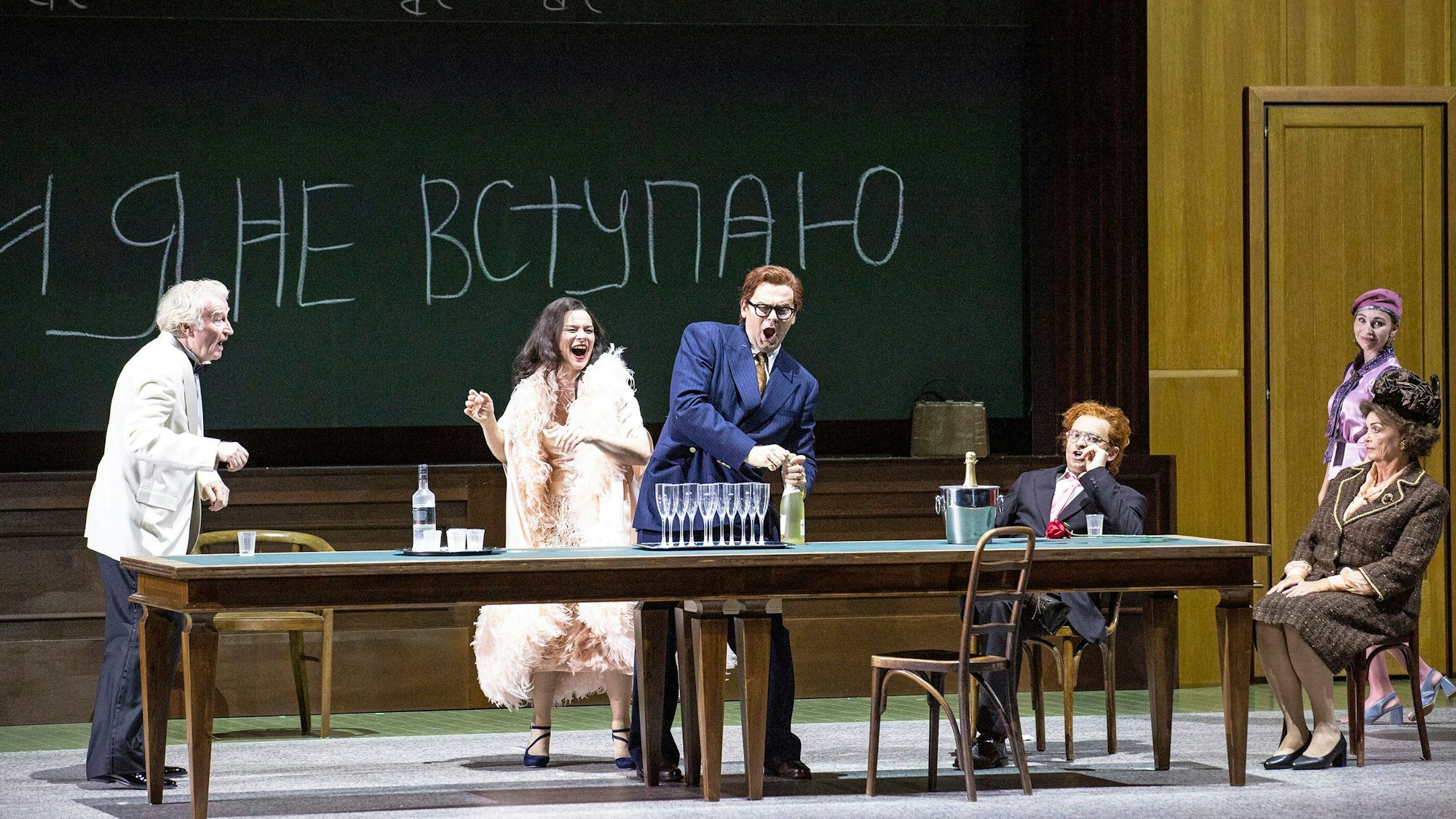 Vor einer beschriebenen Tafel sitzen und stehen Schauspieler um einen langen Tisch herum, während einer eine Flasche Sekt öffnet.
