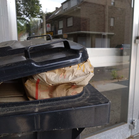 Die Türen des alten Rewe-Gebäudes, davor steht ein Mülleimer mit Fast-Food-Abfällen.