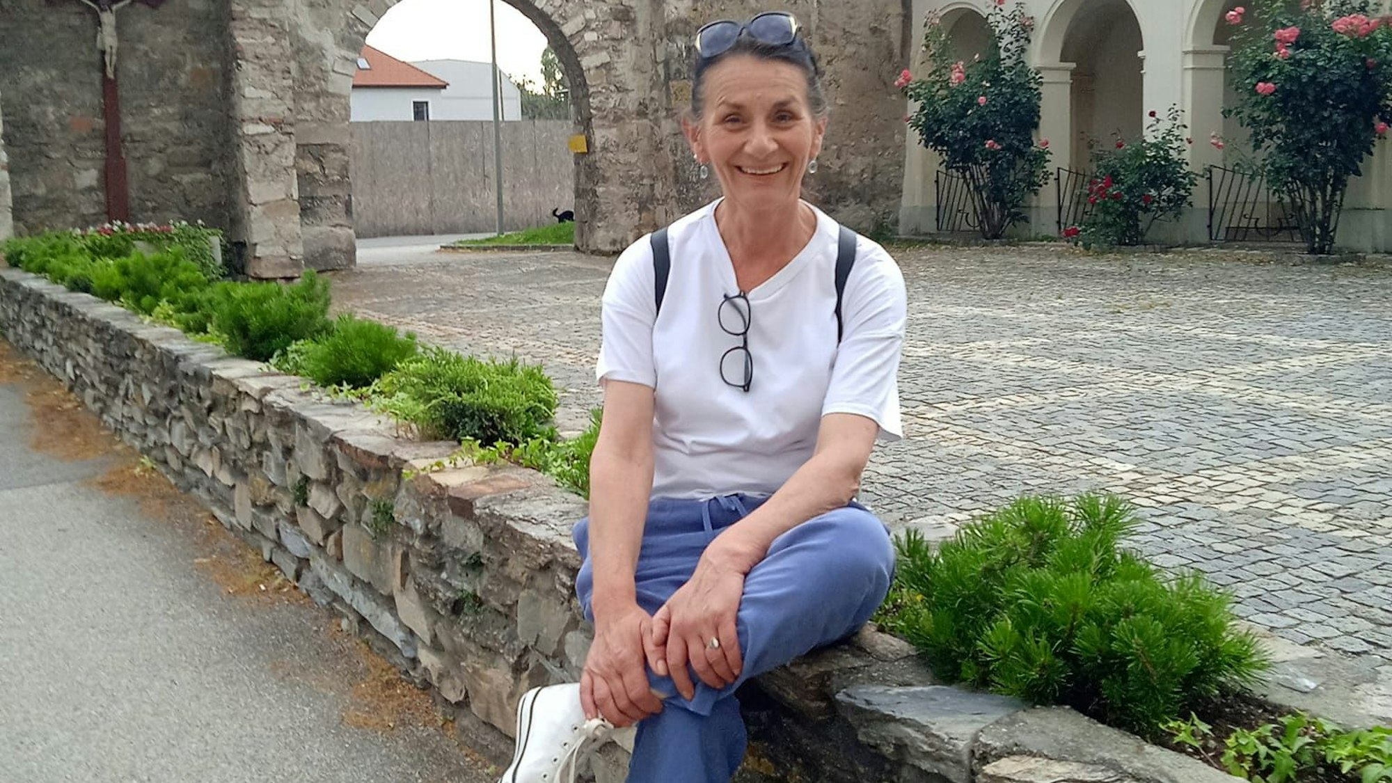 Eine dunkelhaarige Frau sitzt auf einer Mauer aus Stein und lacht in die Kamera.
