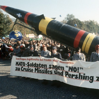 Das Foto zeigt Teilnehmer der Demonstration gegen die Stationierung atomarer amerikanischer Mittelstreckenraketen in Westeuropa am 22.10.1983 im Bonner Hofgarten. Im Vordergrund tragen uniformierte Bundeswehrsoldaten ein Transparent mit der Aufschrift „Nato-Soldaten sagen ‚No!‘ zu Cruise Missiles und Pershing zwo!“ sowie eine Pershing-II-Attrappe. Im Hintergrund sind Massen von Demonstranten mit weiteren, nicht lesbaren Transparenten zu sehen.