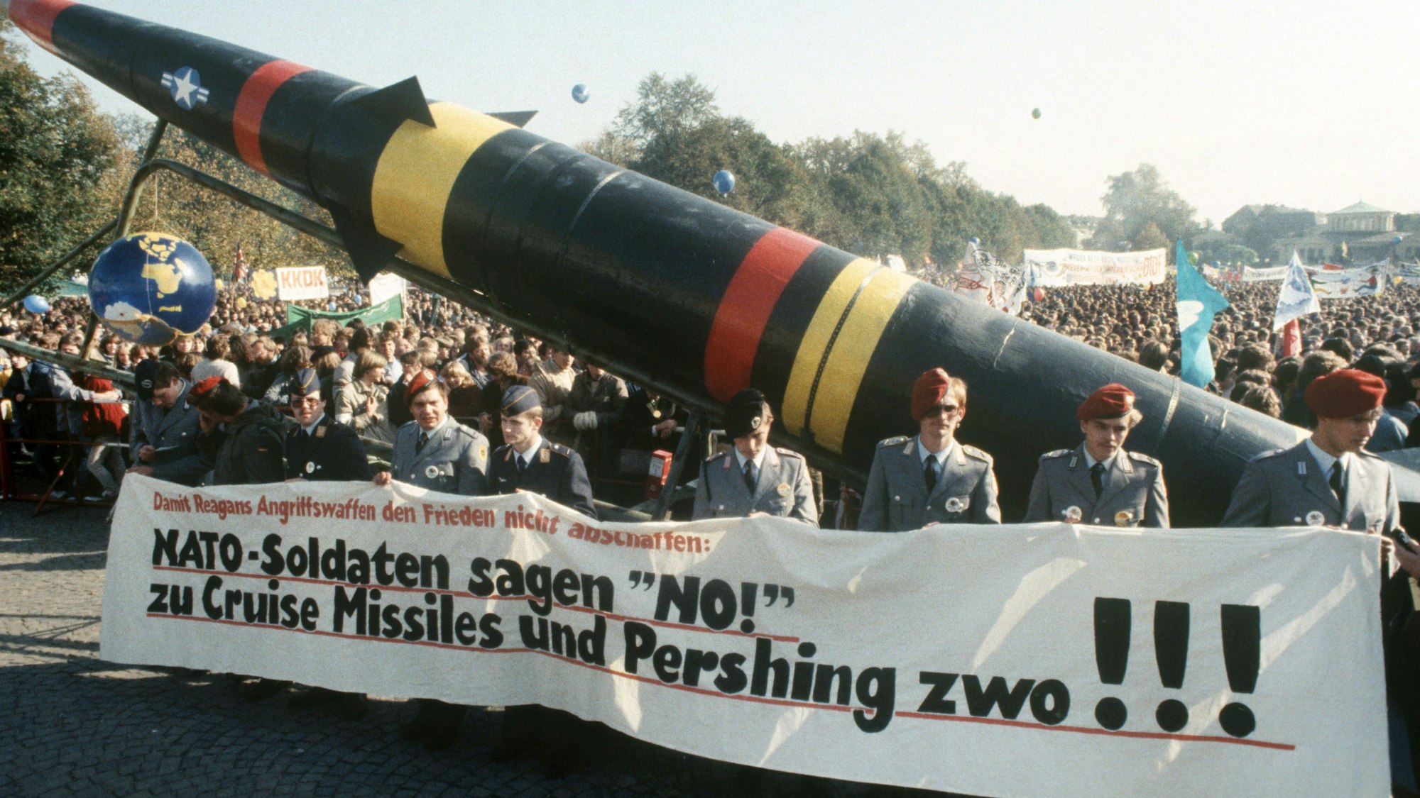 Das Foto zeigt Teilnehmer der Demonstration gegen die Stationierung atomarer amerikanischer Mittelstreckenraketen in Westeuropa am 22.10.1983 im Bonner Hofgarten. Im Vordergrund tragen uniformierte Bundeswehrsoldaten ein Transparent mit der Aufschrift „Nato-Soldaten sagen ‚No!‘ zu Cruise Missiles und Pershing zwo!“ sowie eine Pershing-II-Attrappe. Im Hintergrund sind Massen von Demonstranten mit weiteren, nicht lesbaren Transparenten zu sehen.