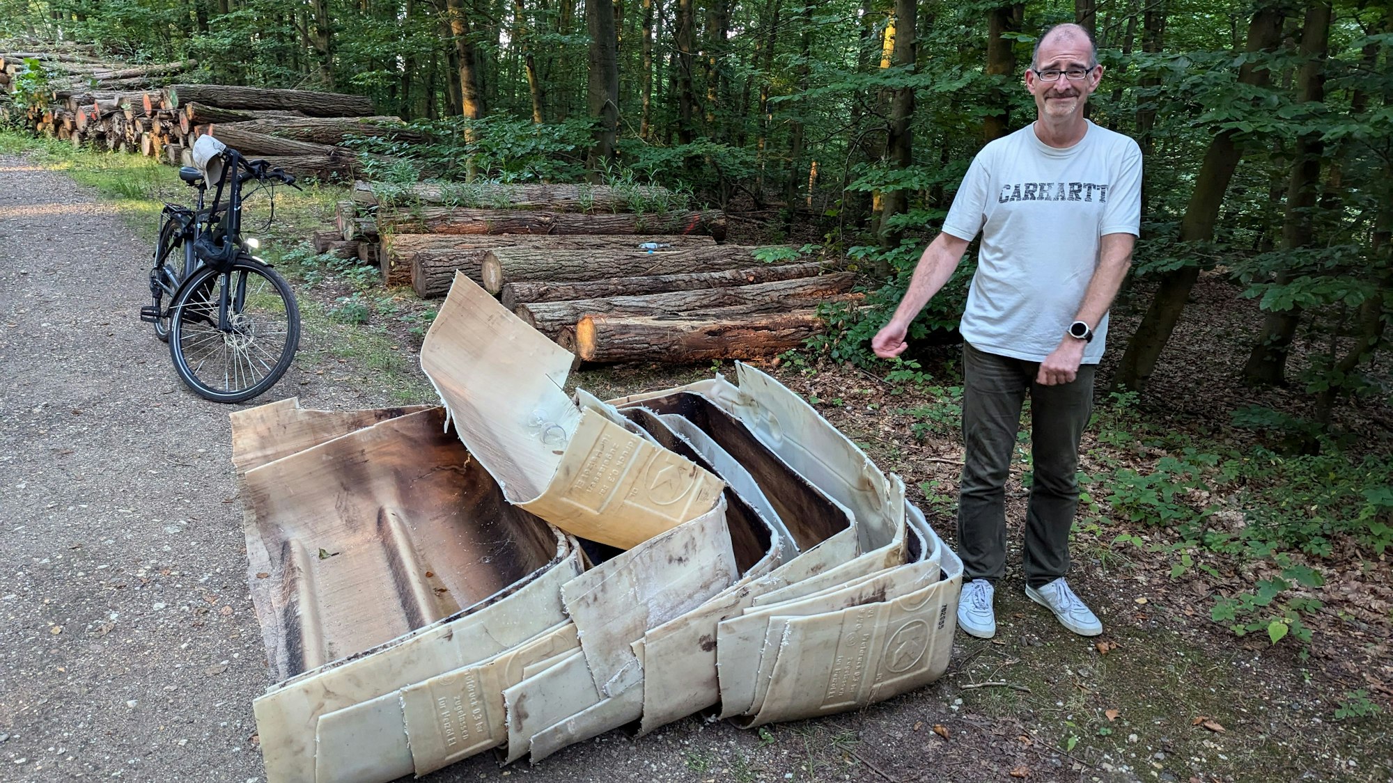 Ein Mann steht neben den zersägten Öltanks vor einem Holzstapel auf einem Waldweg.