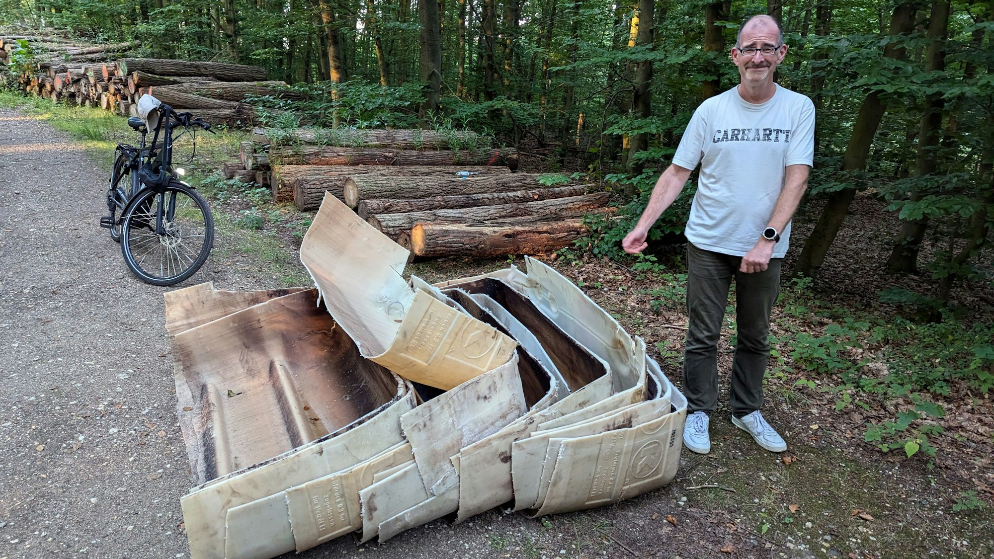 Ein Mann steht neben ineinander verschachtelten alten Kunststoffteilen vor einem Holzstapel auf einem Waldweg.