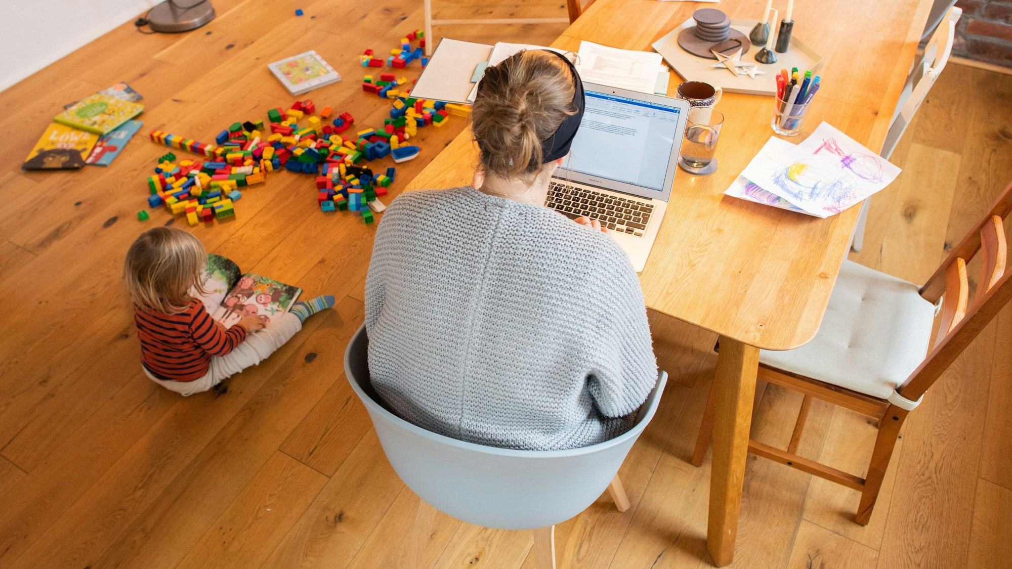 Eine Frau sitzt am Esstisch vor einem Laptop. Am Boden neben ihr spielt ein Kind.
