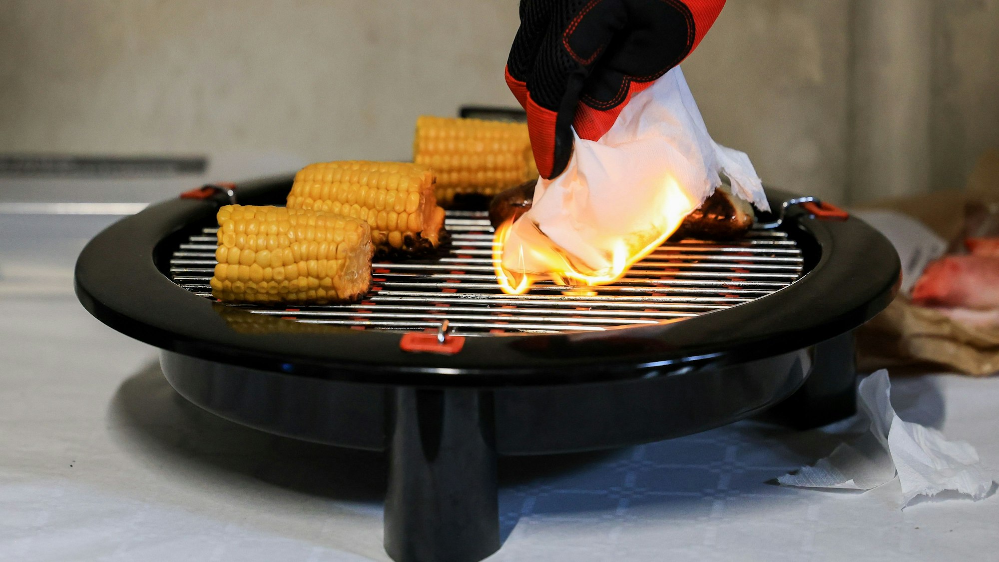 Auf einem Elektrogrill liegen drei kleine Maiskolben, ein Feuerwehrmann entzündet eine Serviette durch bloßen Kontakt mit dem Grill.