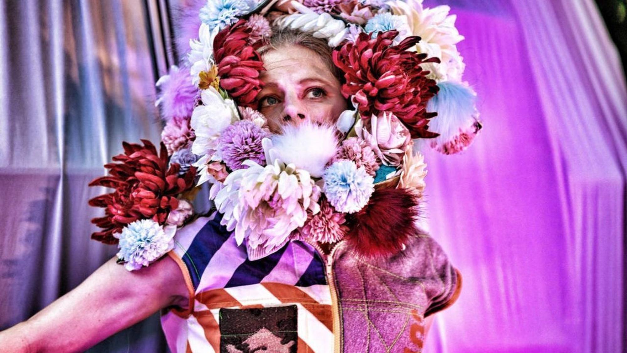 Eine mit einem aufwendigen Blumenkranz geschmückte Schauspielerin vor einer mit Tüchern behangenen Kulisse.