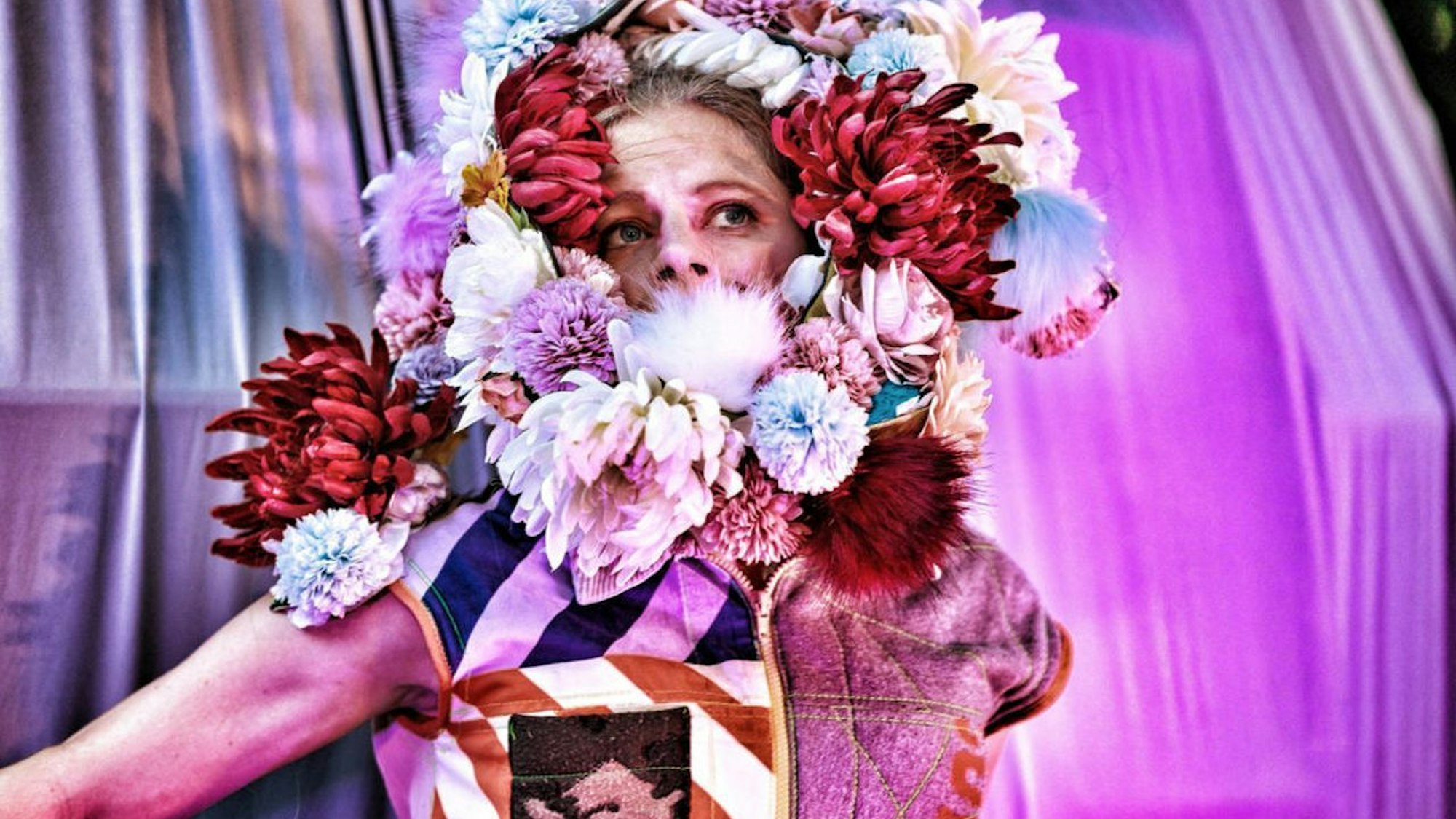 Eine mit einem aufwendigen Blumenkranz geschmückte Schauspielerin vor einer mit Tüchern behangenen Kulisse.