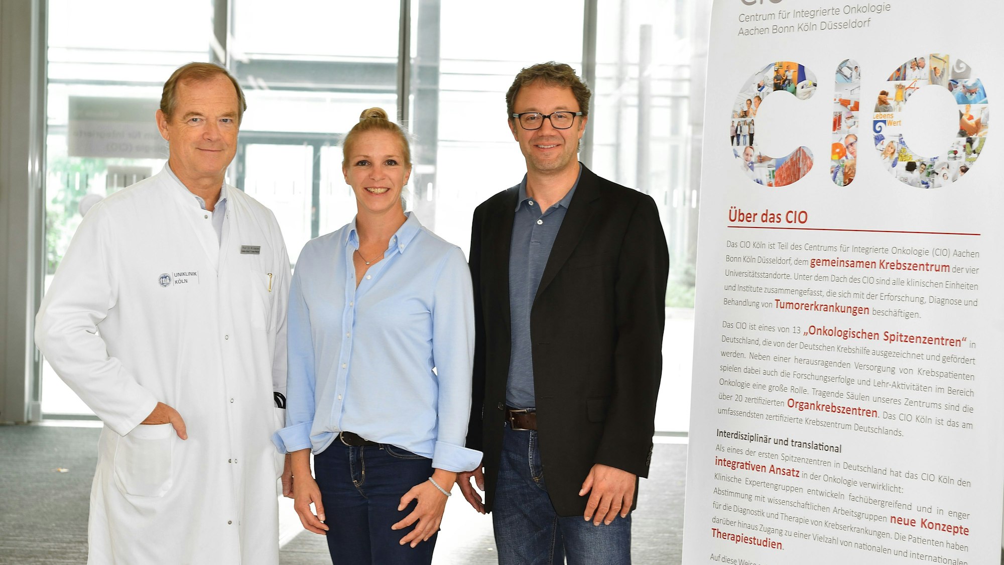 Zwei Männer stehen links und rechts von einer Frau. Neben ihnen steht ein Aufsteller mit der Aufschrift CIO und einem Text darüber