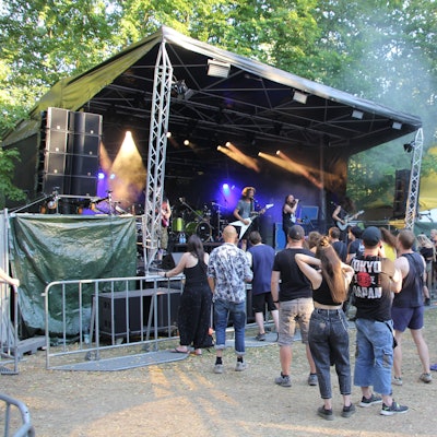 Auf dem Bild ist die Bühne in Frechen zu sehen, eine Band tritt gerade auf. Vor der Bühne steht eine Menschenmenge. Im Hintergrund einige Bäume.