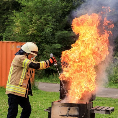 Ein Feuerwehrmann spritzt Wasser aus einer Flasche auf einen Fettbrand und erzeugt damit eine große Stichflamme.