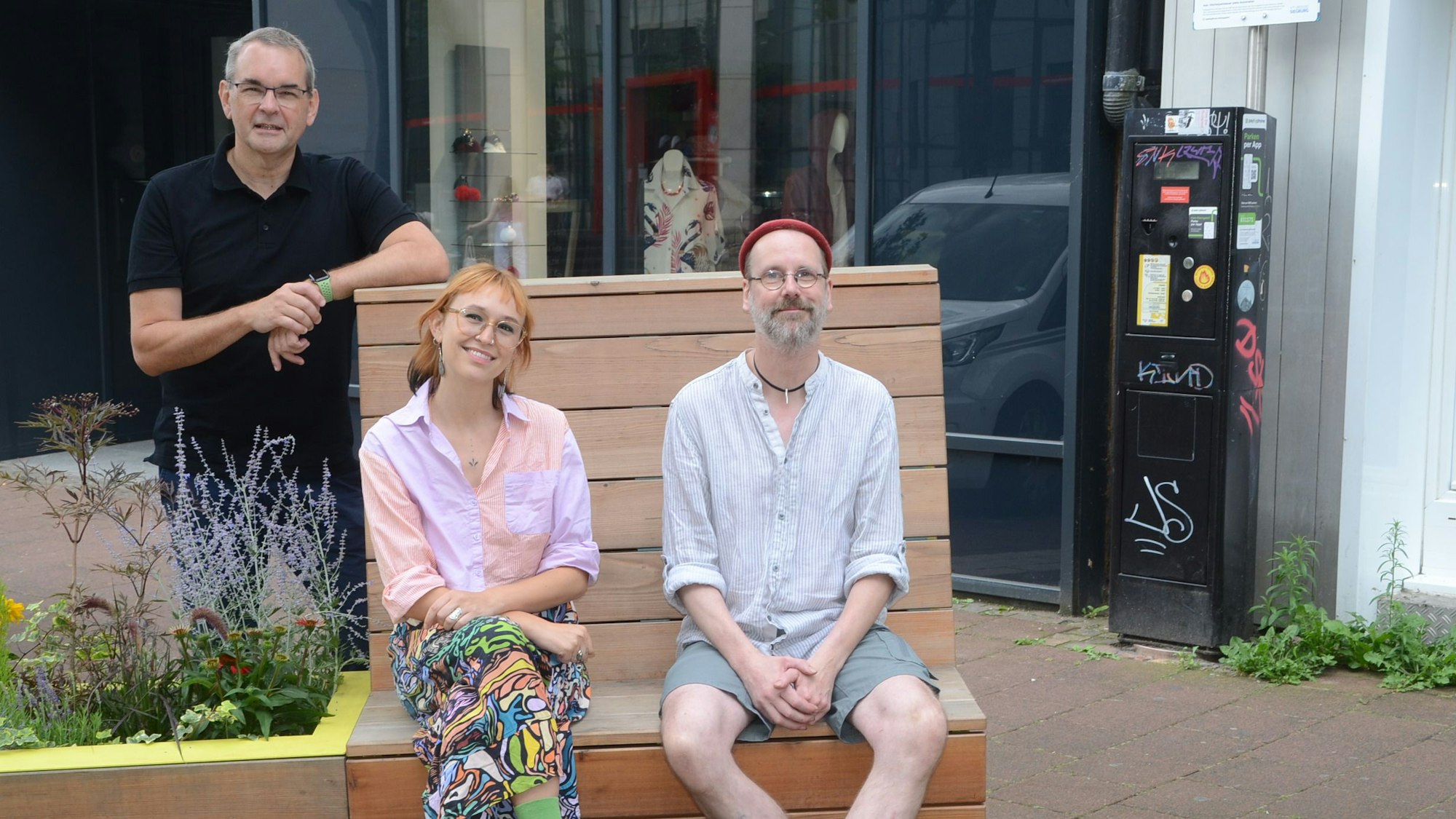 Ein Mann und eine Frau sitzen auf einer Bank, in die ein Blumenkübel integriert ist. Hinter der Bank steht ein Mann im schwarzen Polohemd.