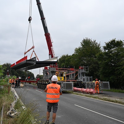 Ein Bauarbeiter in orangefarbener Warnweste beobachtet, wie das tonnenschwere Stahlbrückenteil mit dem Schwerlastkran verladen wird.