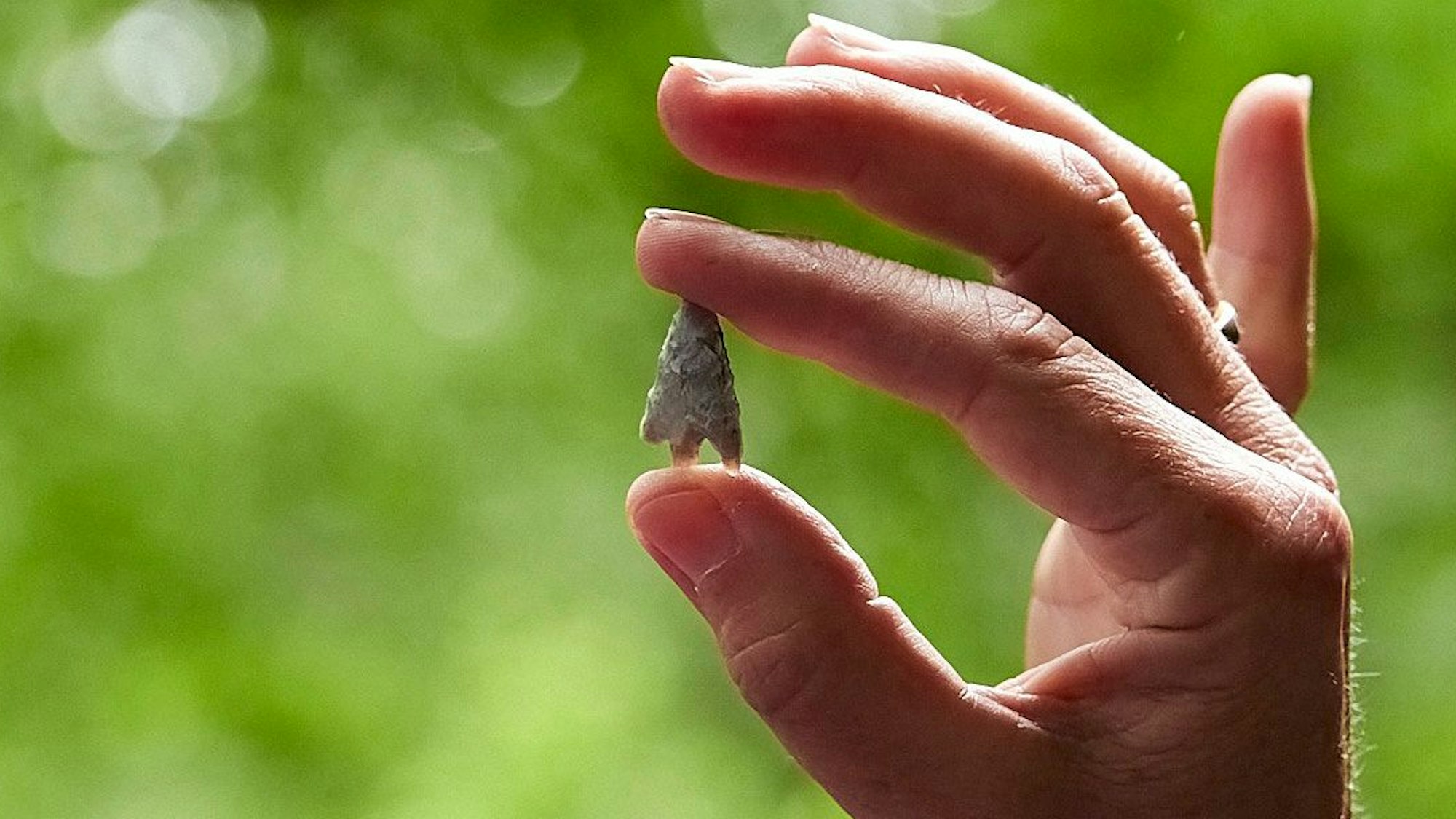 Eine Frauenhand ist vor einem grünen Hintergrund zu sehen. Zwischen Daumen und Zeigefinger hält sie eine Pfeilspitze aus der Steinzeit.