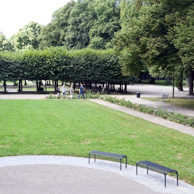 Zu sehen ist eine Grünfläche mit Sitzbänken, Kieswegen, Lindenbäumen und Blumenbeeten.