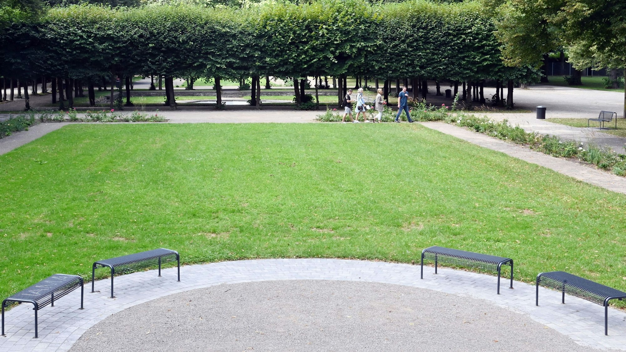 Zu sehen ist eine Grünfläche mit Sitzbänken, Kieswegen, Lindenbäumen und Blumenbeeten.