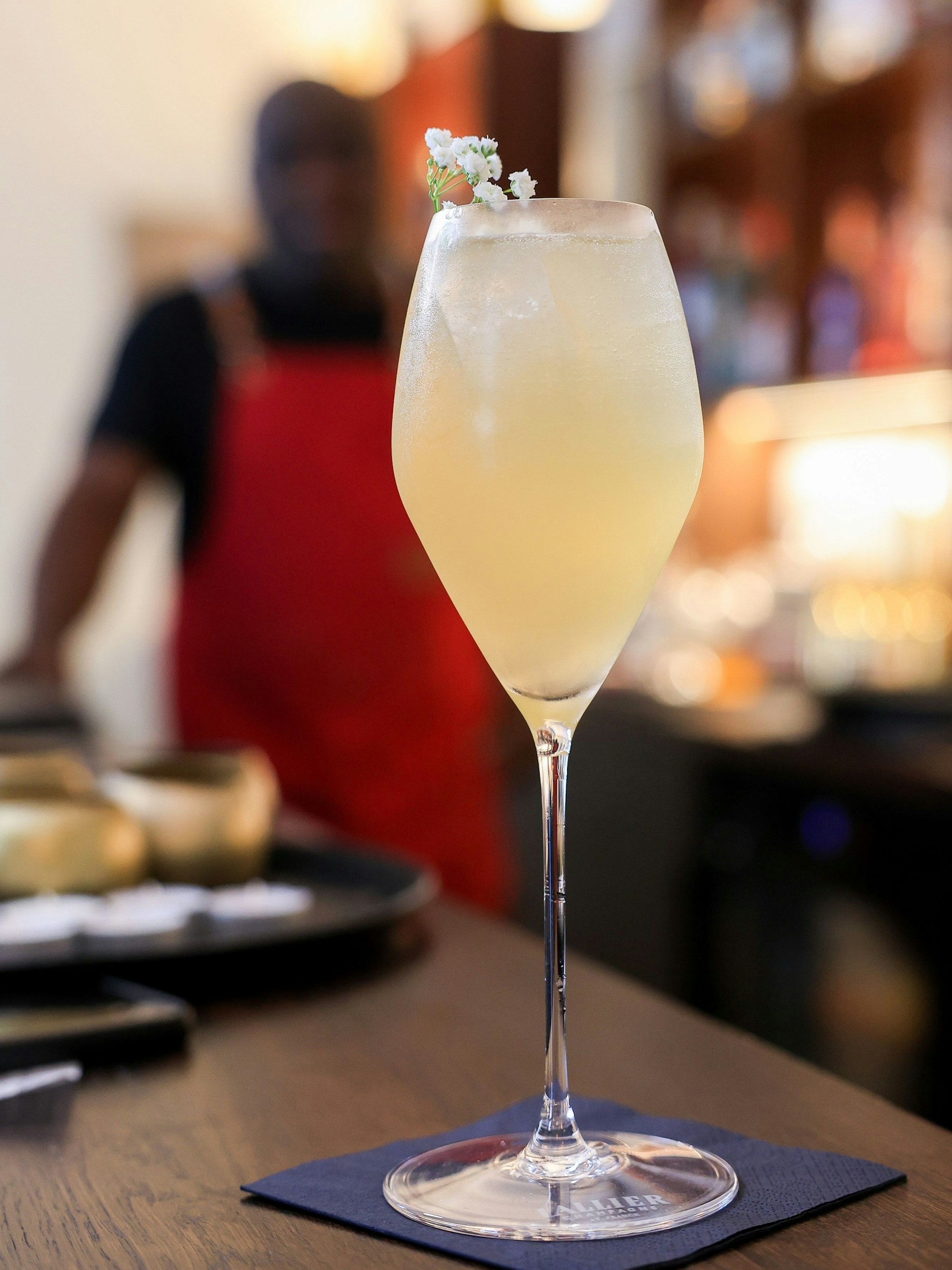 Ein Champagnerglas steht auf dem Tresen und ist mit einer kleinen weißen Blüte verziert. Im Hintergrund sieht man verschwommen den Barchef stehen.