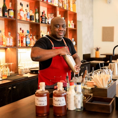 Barchef Samuél Antonio Bernardo steht an der Bar und hat einen Shaker in der Hand. Er lächelt in die Kamera.